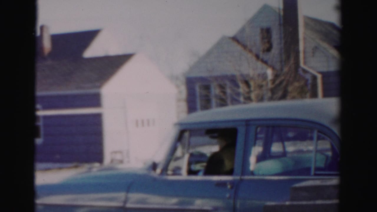 Blue sedan parked in front of houses with a man seated at the steering wheel.