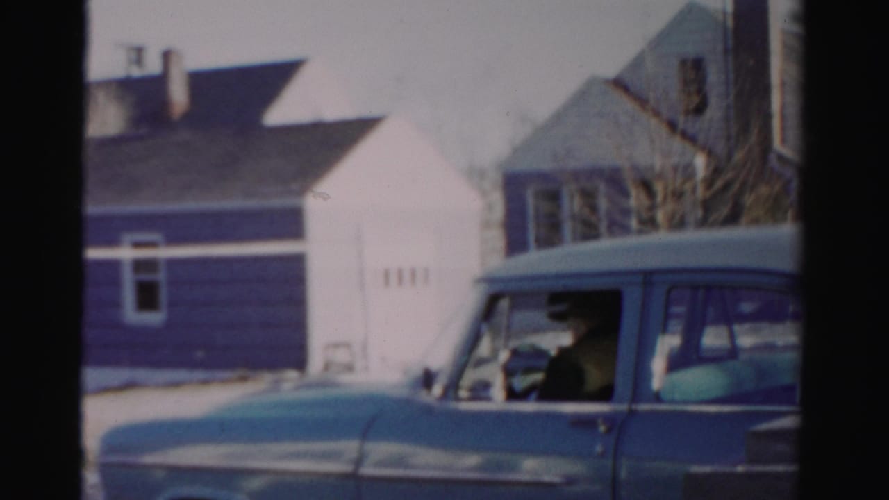 Man at the wheel of a blue sedan with blue and white houses behind him.