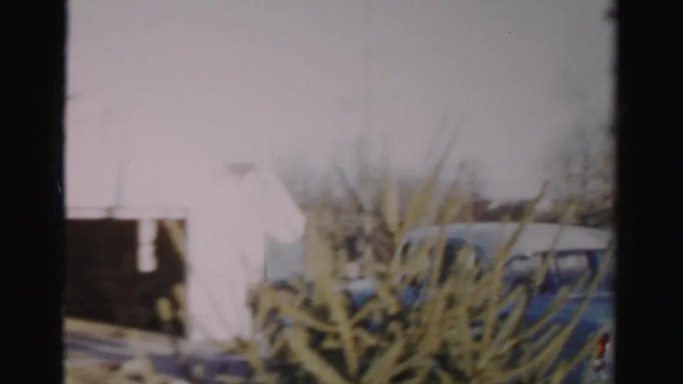 1950s Albany home-movie frame showing a blue sedan near a white garage-like building and evergreen tree.