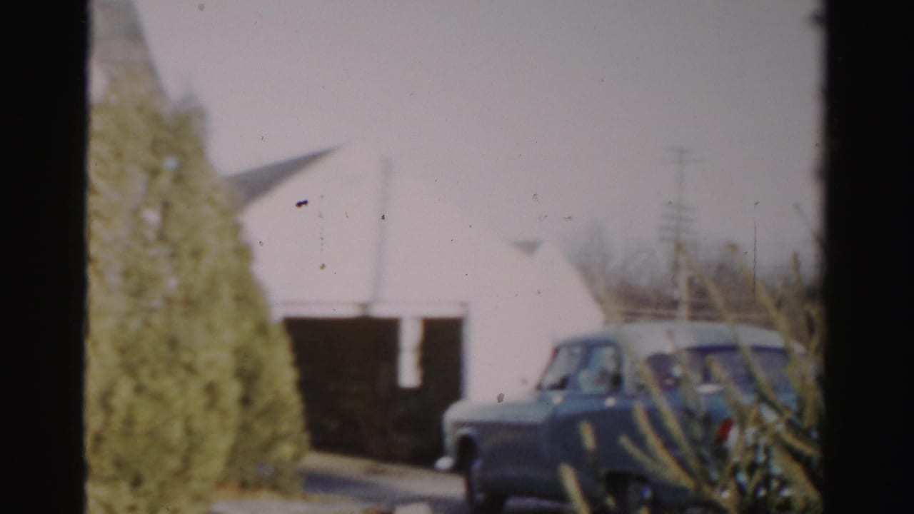 Blue car near a white building with shrubs in the foreground.