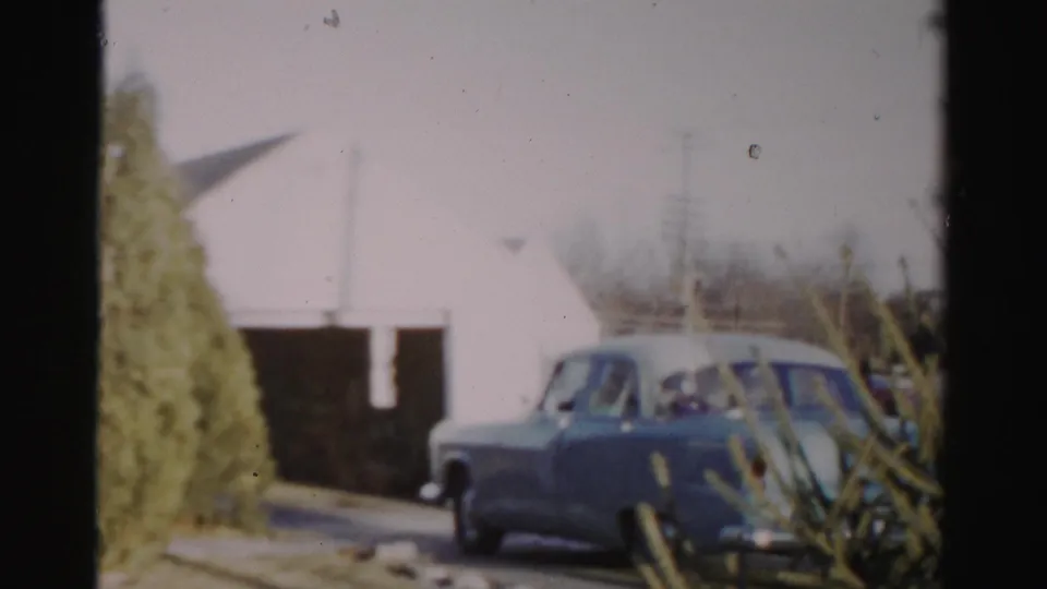 Albany-area 1950s home-movie frame with a blue sedan near a driveway and white garage-like building.