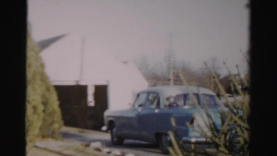 Blue sedan near a white garage-like building, with bare branches and 8mm film texture visible.