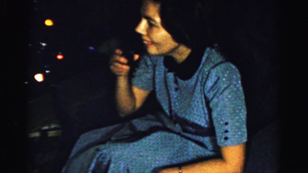 In metadata-supported Albany, 1956, an adult woman in a blue patterned dress sits in low light with small out-of-focus lights nearby.