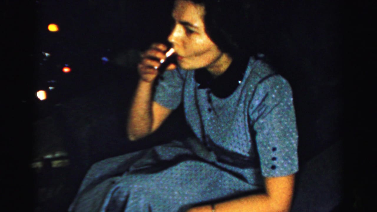 Adult woman in a blue dress raises a cigarette or small object near her mouth in darkness.