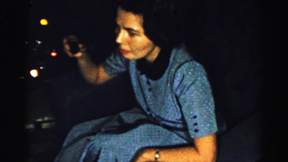 The 1956 Albany home-movie frame shows the seated woman turned sideways, holding a small glass or object in the dim light.