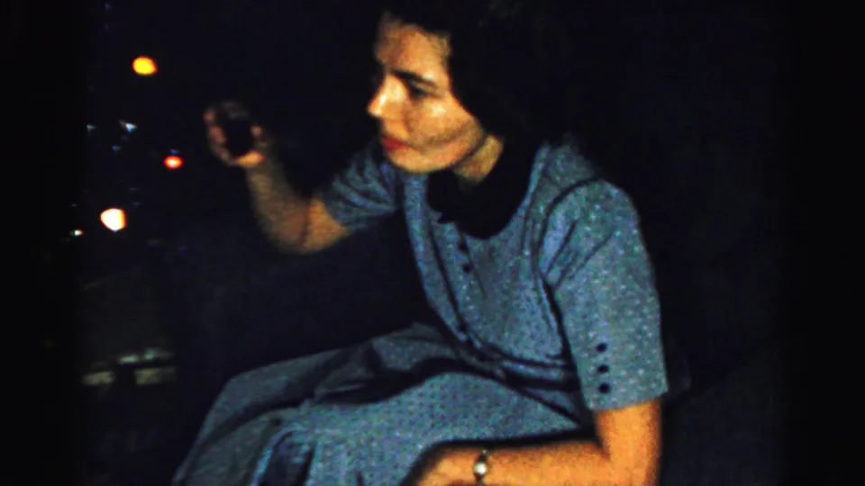 In this metadata-supported Albany 1956 frame, the woman remains seated in a blue dress, partly lit against a dark background with distant lights.