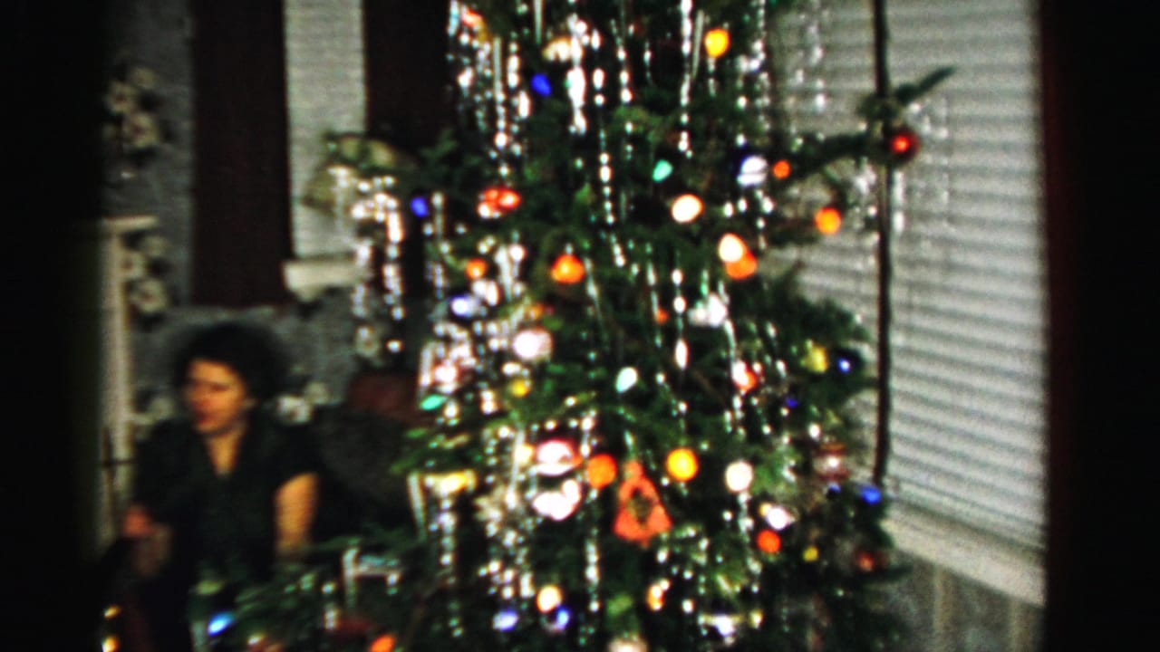 In Albany home-movie footage from 1956, a woman sits near a Christmas tree with colored lights, tinsel, blinds, and patterned carpet.