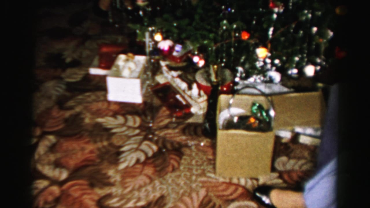 Albany 1956 home-movie frame showing a decorated tree, boxes and gift items on a patterned carpet in a private interior.