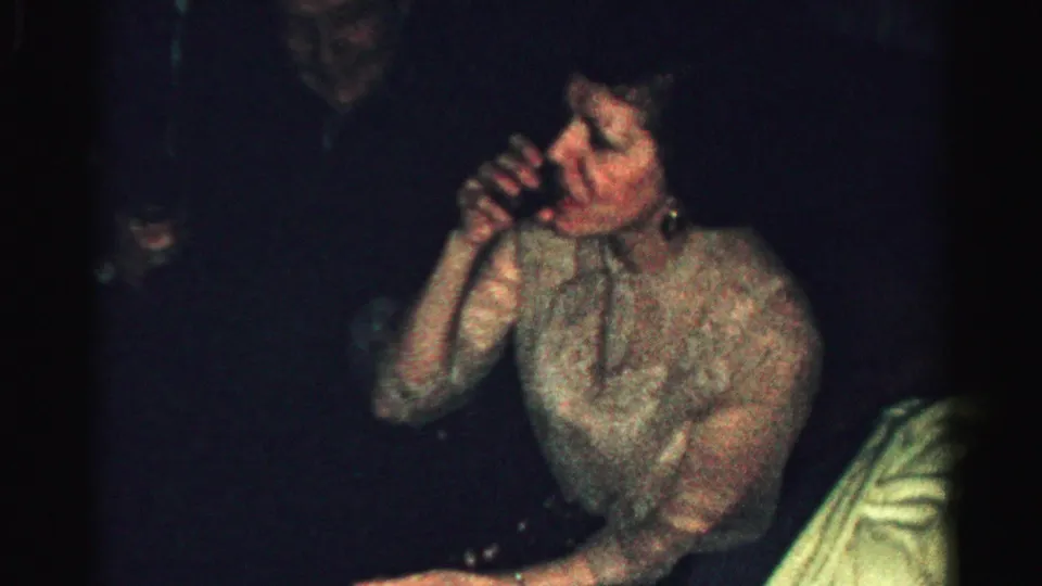 Albany 1956 home-movie frame showing an adult woman in a light textured dress sipping from a small glass in a private room.