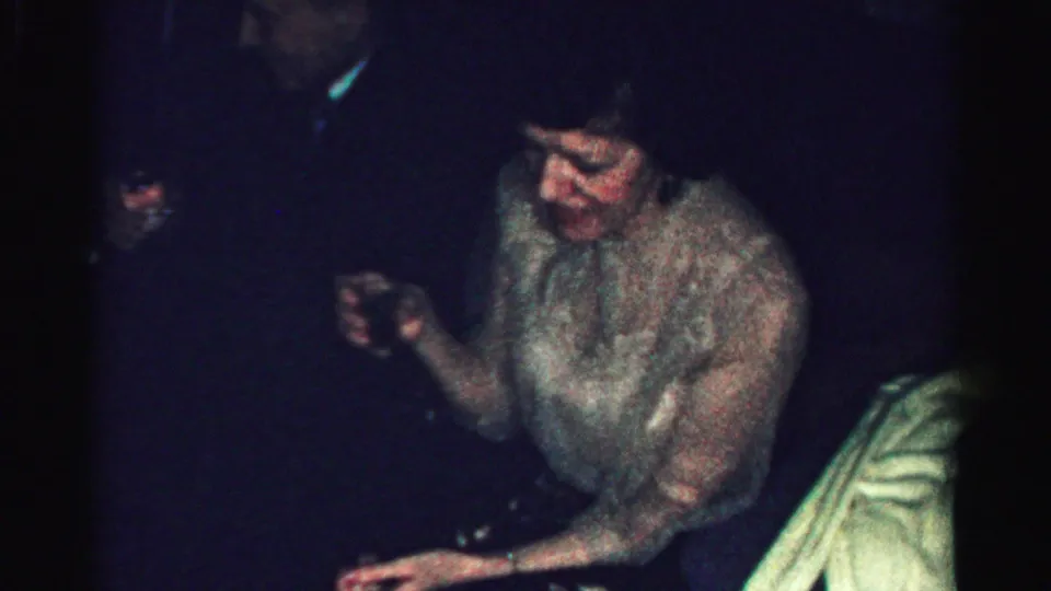 Albany 1956 home-movie frame of a seated adult woman holding a drink and looking down in a low-lit private interior.