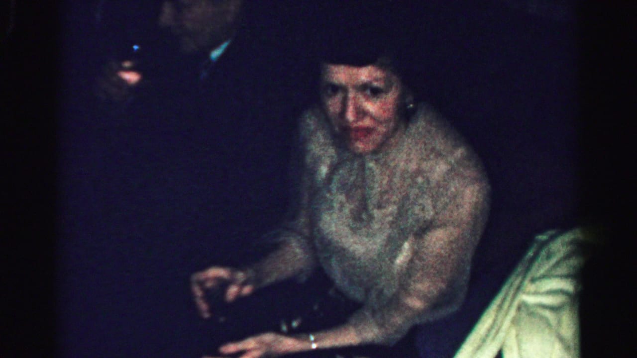 Adult woman at a dim Albany, New York private party, seated in a light textured dress during 1950s home-movie footage.
