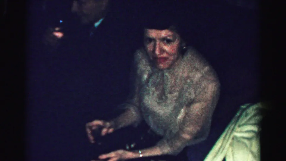 Adult woman at a dim Albany, New York private party, seated in a light textured dress during 1950s home-movie footage.