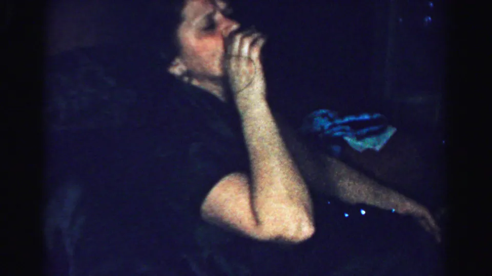 Adult woman raises a glass to drink in a dim Albany, New York private-party scene from 1950s 8mm footage.