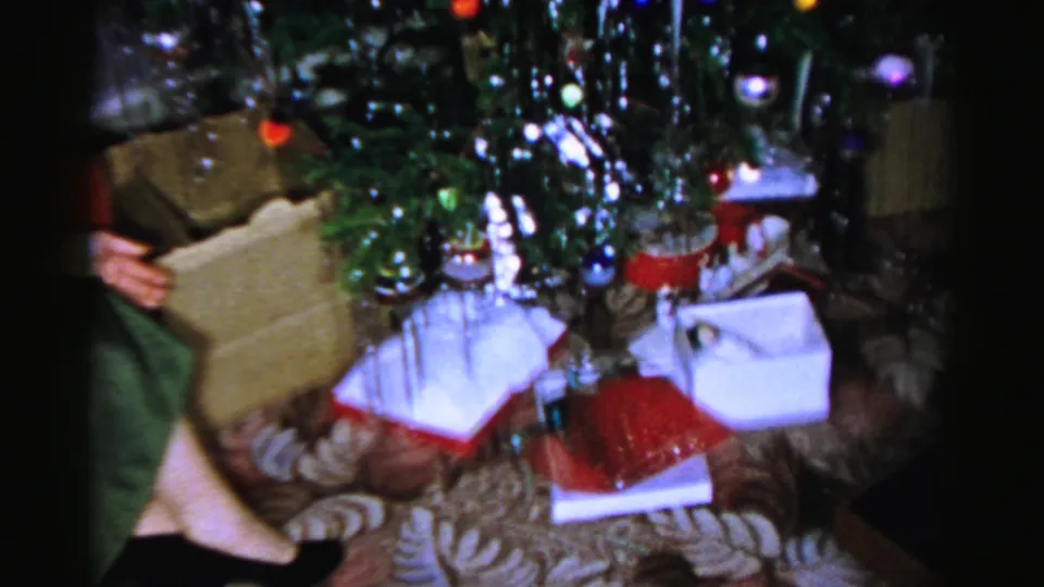Wrapped boxes and bags are clustered under the Christmas tree, with a person's hand partly visible at the side.