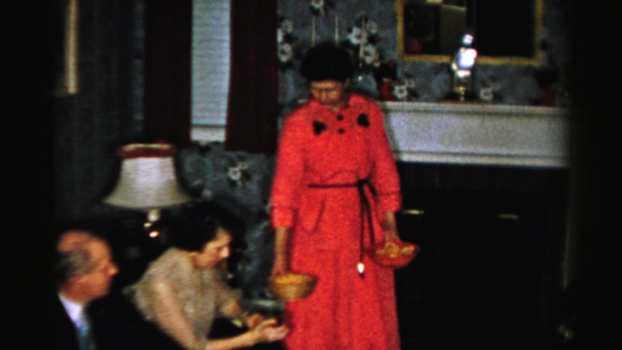 In a 1956 Albany home-movie frame, a woman in a red dress and black hat serves food near seated adults in a decorated living room.