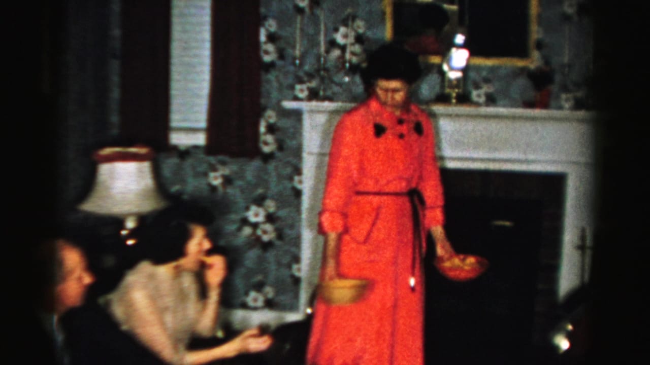 Woman in red dress stands by seated guests with bowls in a living room.