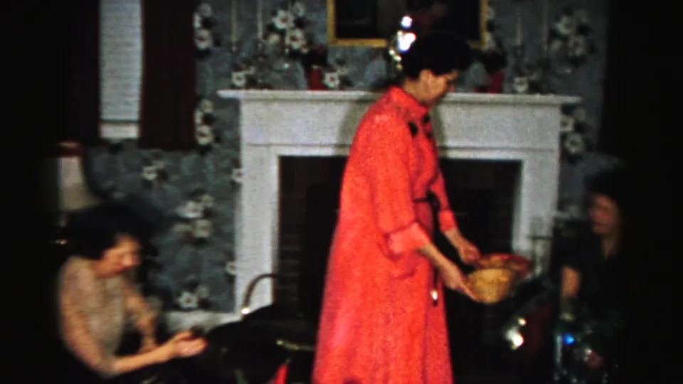 The woman in red leans toward guests with a bowl in an Albany living room, with the fireplace and holiday decor behind her.