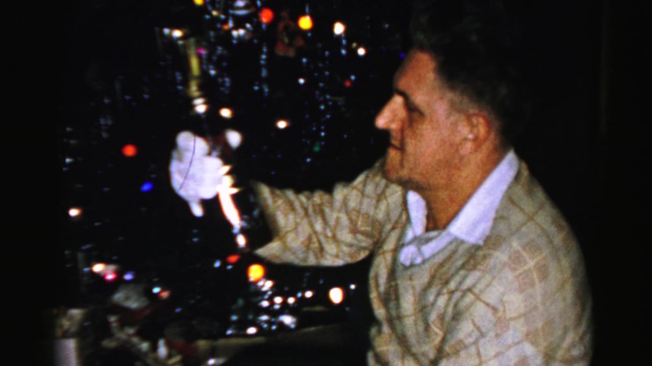 Man in a patterned sweater and white gloves holds up a dark bottle near a Christmas tree.