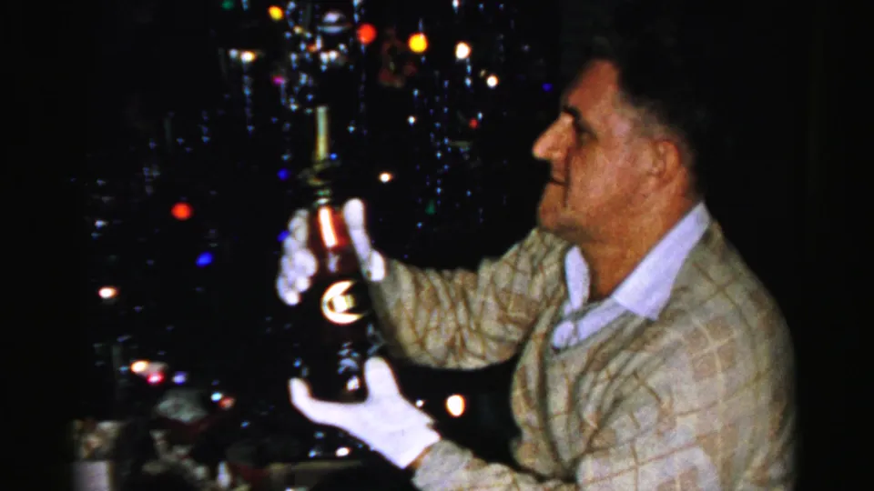 The man turns slightly while holding a dark bottle with white gloves, framed by tinsel, colored lights, and a dim holiday interior.