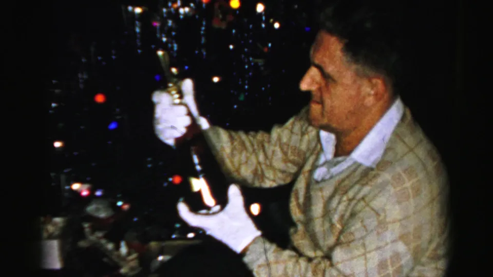 A smiling man in a light patterned sweater holds up a bottle with gloved hands near the Christmas tree in the private Albany living room.