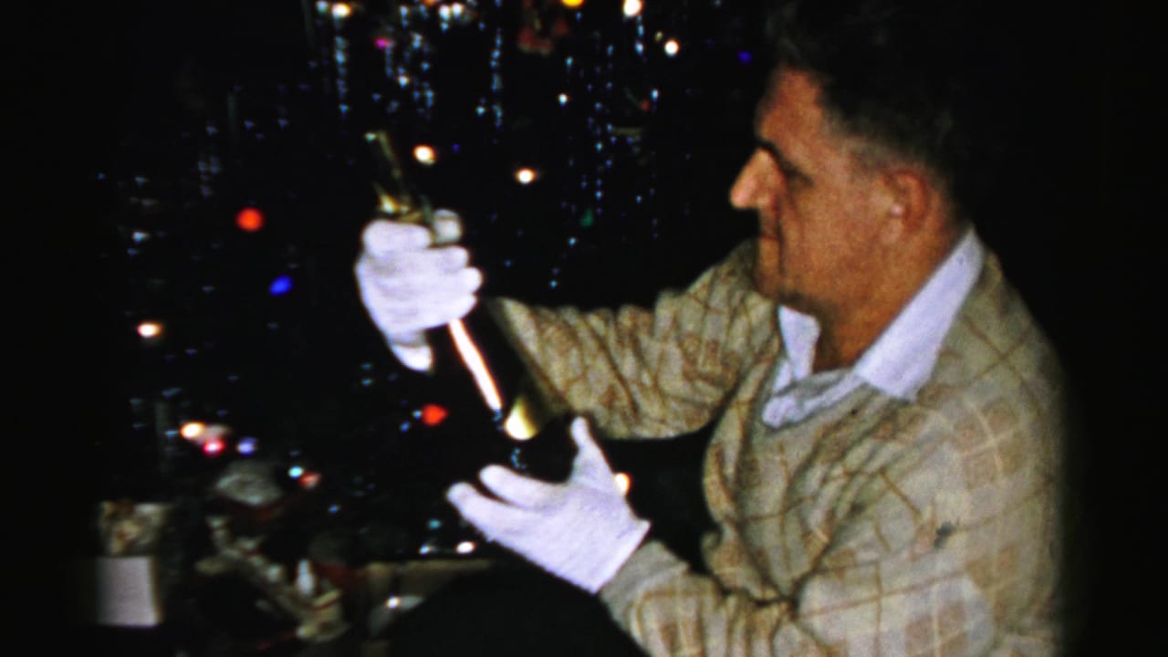 Man in a patterned sweater and white gloves holds a bottle beside a lit Christmas tree.