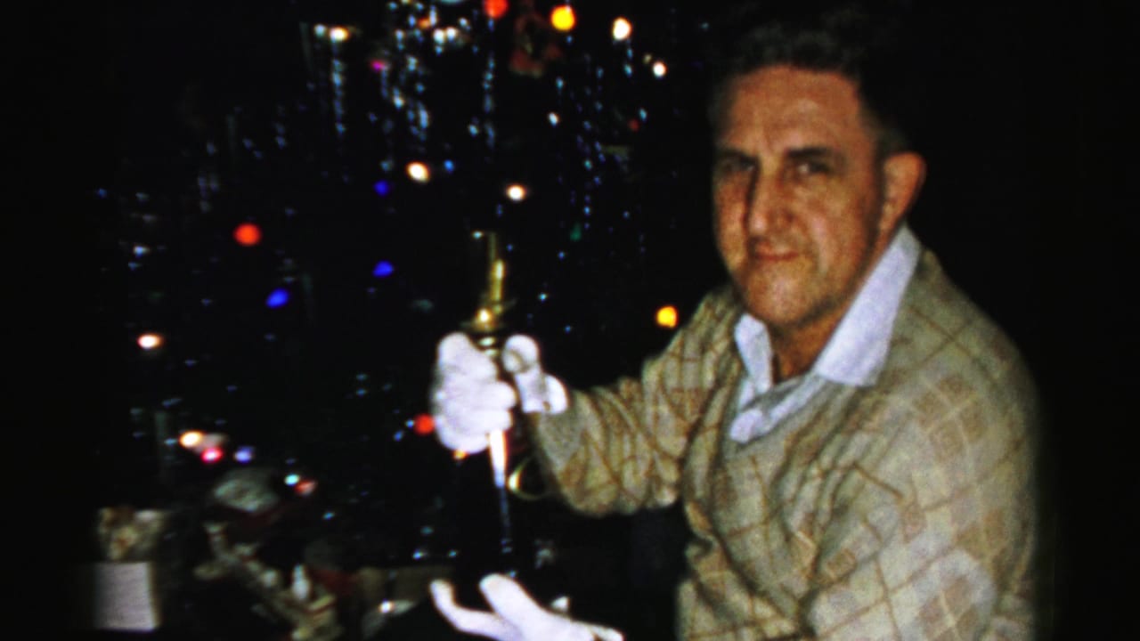 Man faces the camera while holding a bottle in front of a dark, lit Christmas tree.