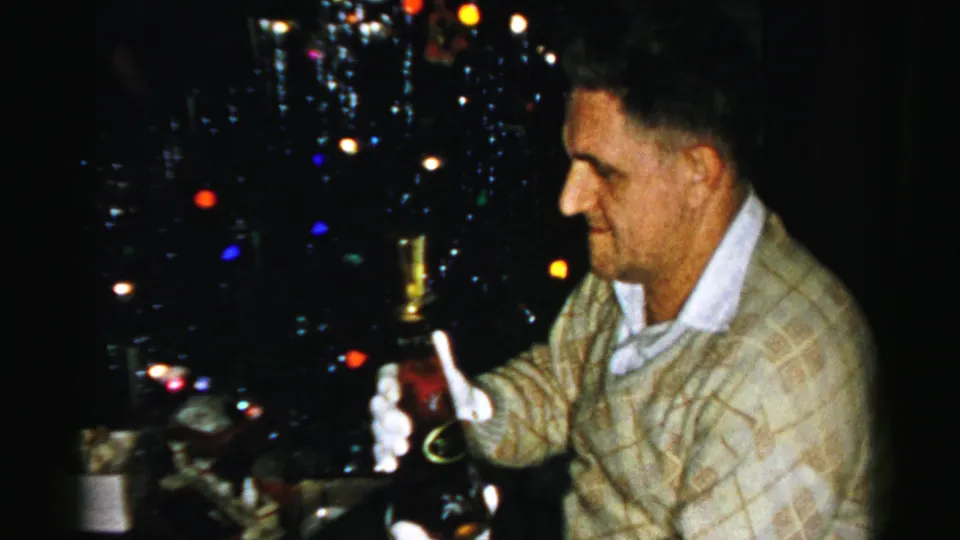 The man lowers the bottle and appears to smile, framed by multicolored Christmas lights in a private holiday interior.