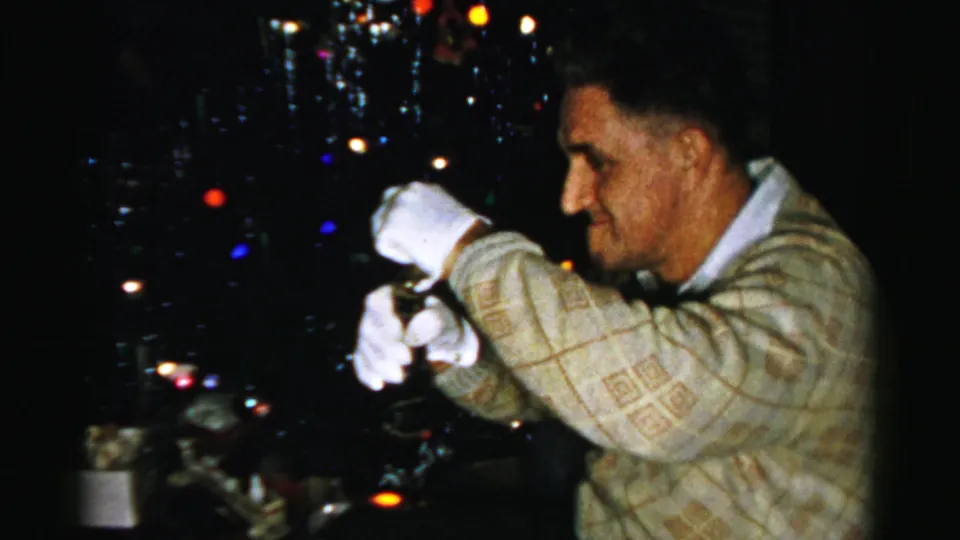The man uses both white-gloved hands on the bottle, appearing to prepare or open it beside the Christmas tree lights.