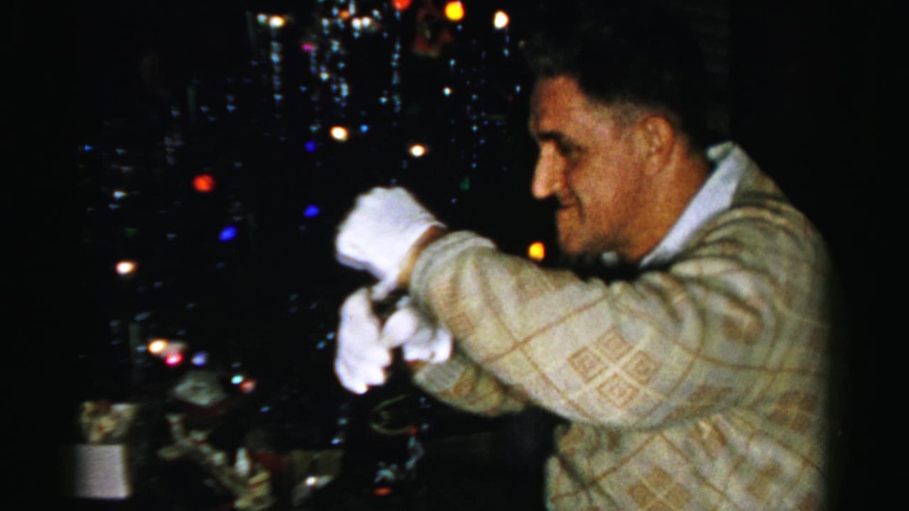 In Albany home-movie footage from 1956, a man in white gloves works at a bottle near a dark, colorfully lit Christmas tree.