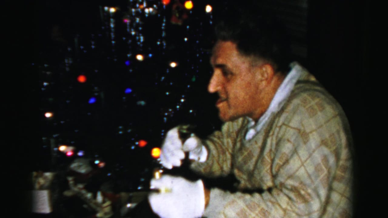 Man in white gloves holds a bottle and cork near a brightly lit Christmas tree.