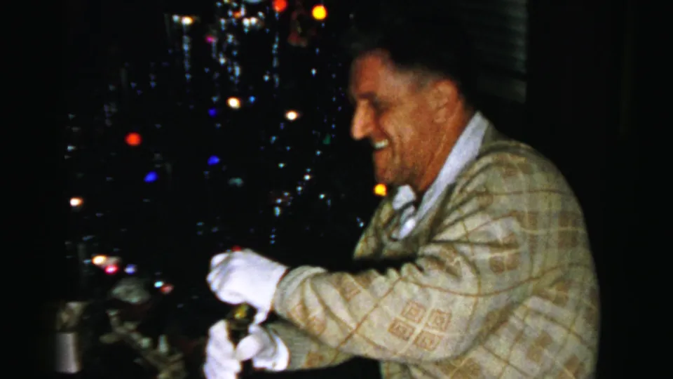 A smiling man in a patterned sweater and white gloves holds the bottle, framed against the dim Christmas tree lights.