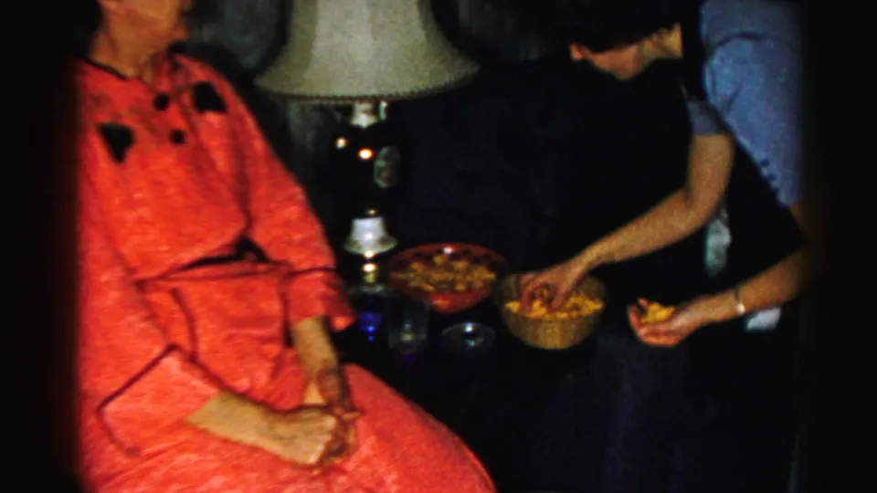A woman in a red dress sits in the private holiday interior while another person leans over bowls of snacks near a table lamp.