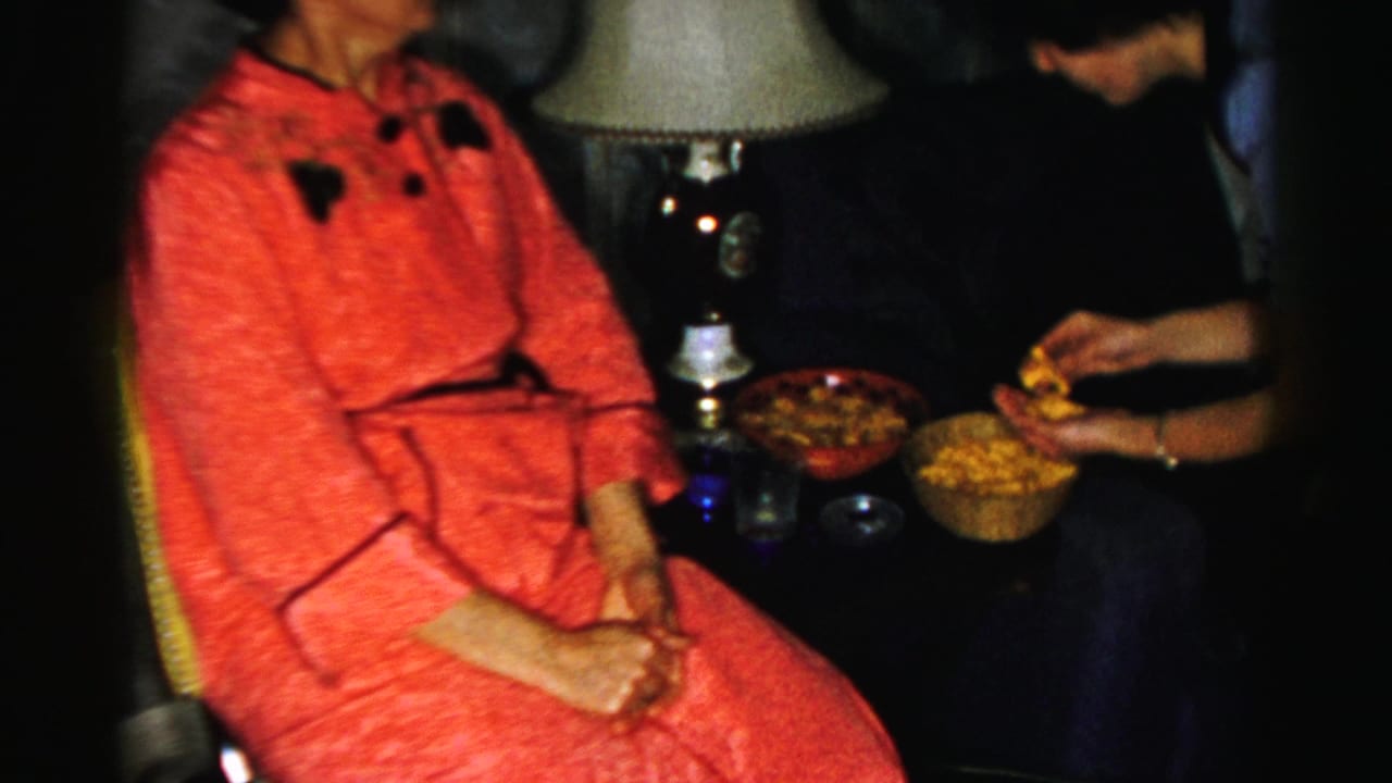 Seated woman in an orange dress beside snack bowls as a partially visible person handles food.