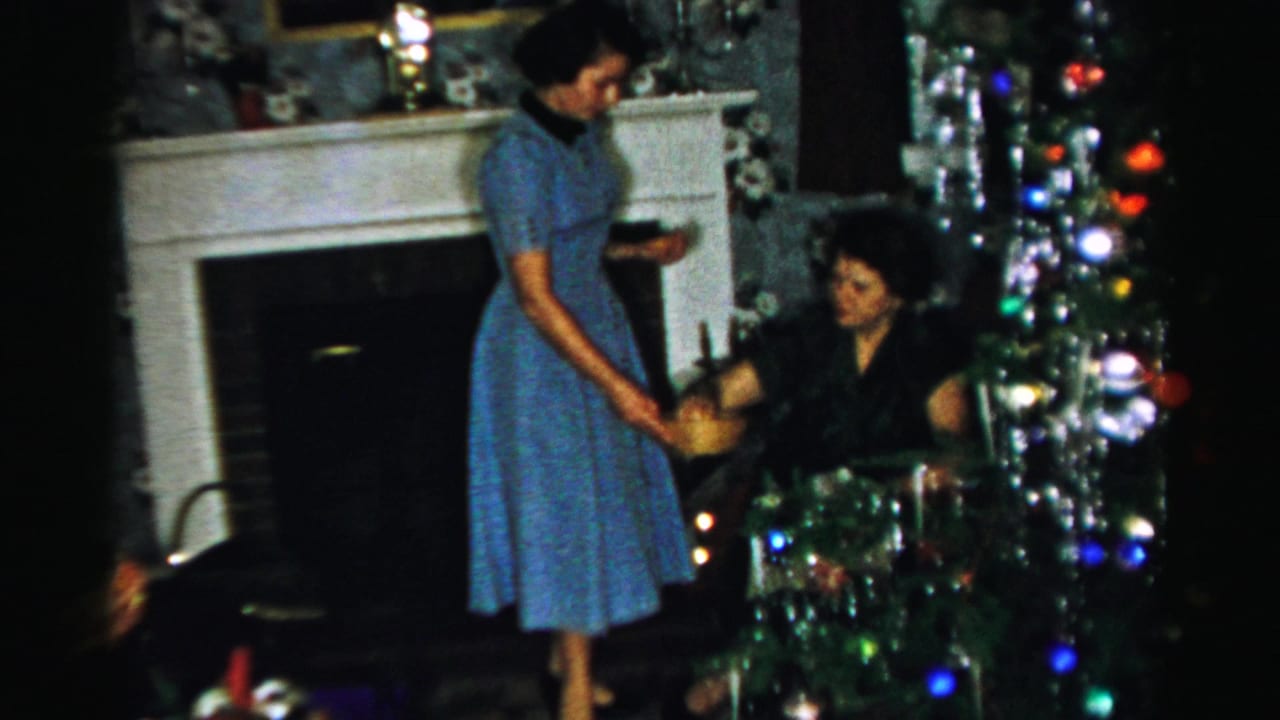 In a mid-1950s Albany home interior, a woman in a blue dress offers a bowl while another woman sits near the Christmas tree.