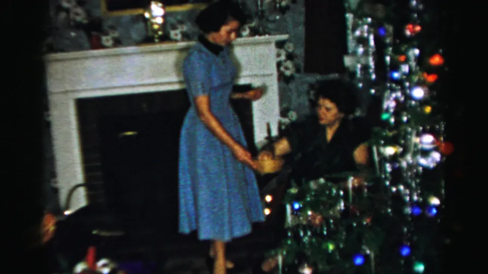 In a mid-1950s Albany home interior, a woman in a blue dress offers a bowl while another woman sits near the Christmas tree.