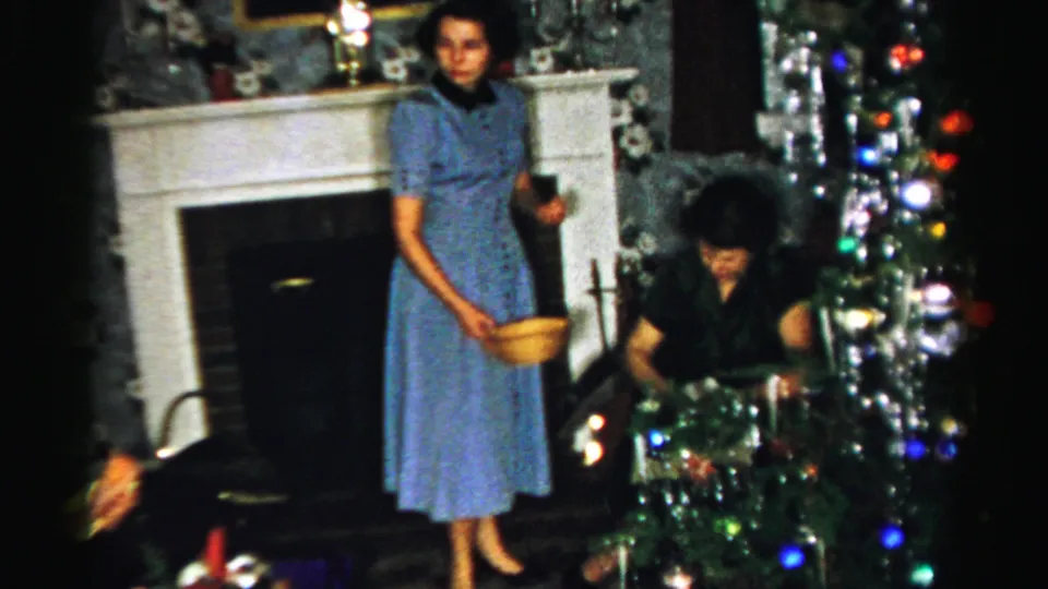 A woman in a blue dress stands by the fireplace and Christmas tree, holding a yellow serving bowl in a private home interior.