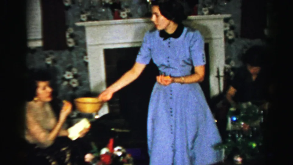 Inside the Albany holiday gathering, a woman in a blue dress passes a yellow bowl to a seated guest while another person remains near the tree.