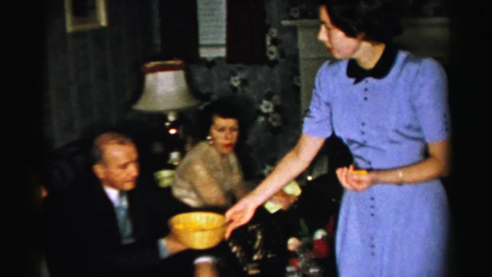 A woman in blue serves a yellow bowl to seated adults near a lamp, fireplace, and decorated Christmas tree in the Albany home scene.