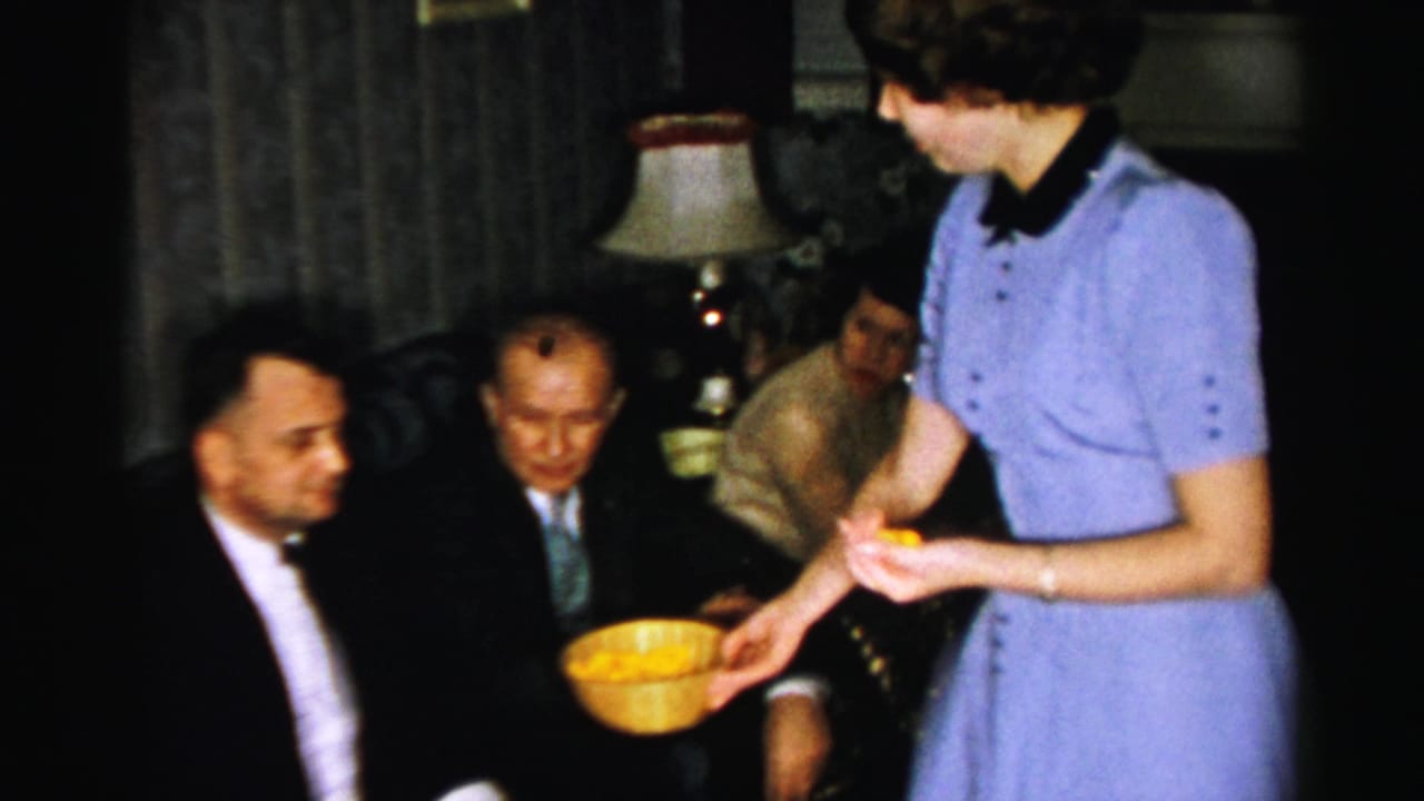 In a 1950s Albany living room, a woman in a blue dress serves snacks from a yellow bowl to men seated in suits.