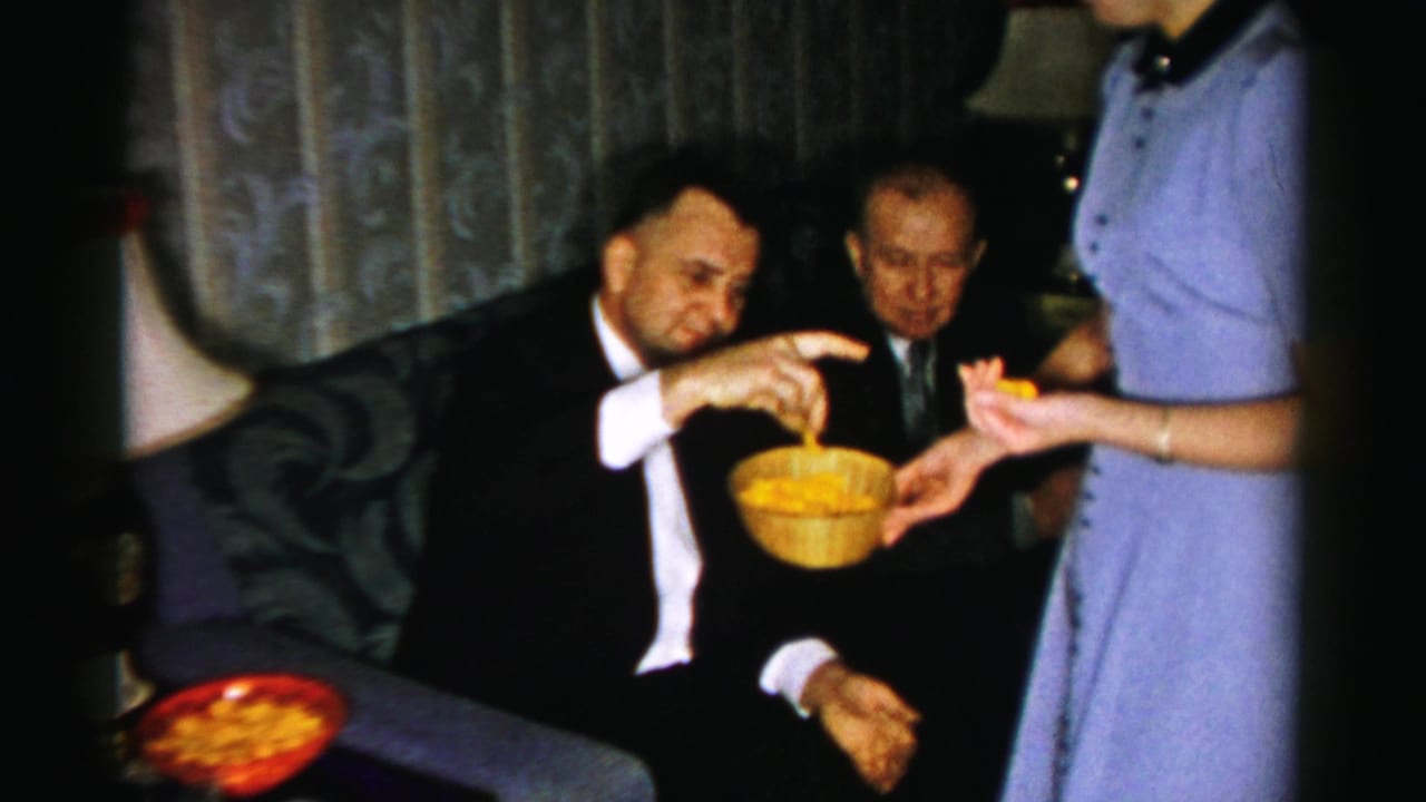 Seated man reaches toward a yellow snack bowl held by a woman in blue.