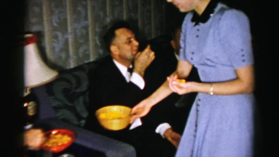 A man in a dark suit eats from his hand while the woman in a blue dress holds the yellow bowl in a softly lit living room.