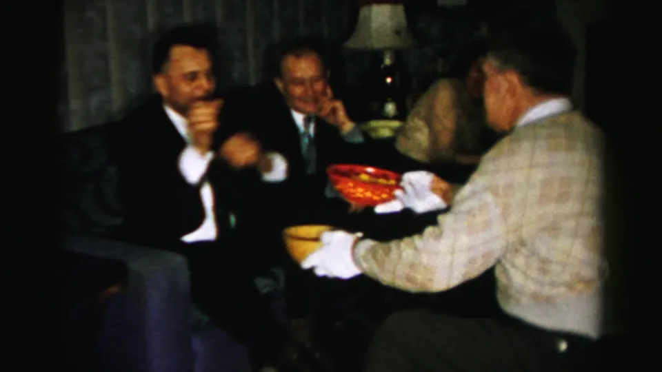 Adults in suits and dresses gather closely in a private Albany living room with yellow and red snack bowls visible between them.