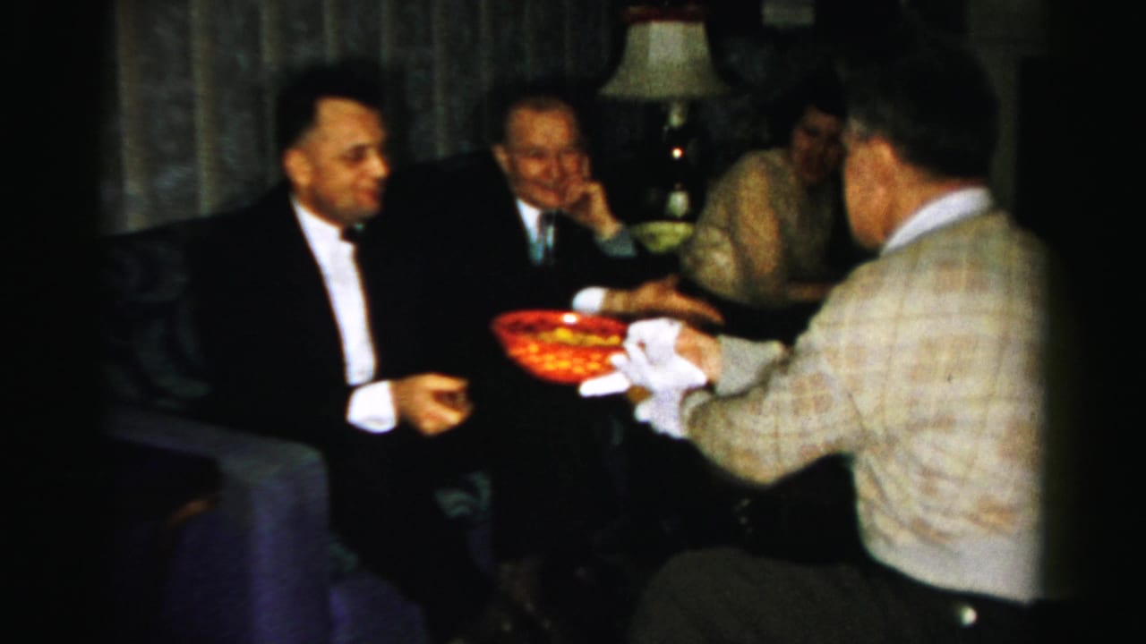 Albany, New York home-movie frame from 1956 showing a man in a patterned sweater offering a red snack bowl to seated party guests.