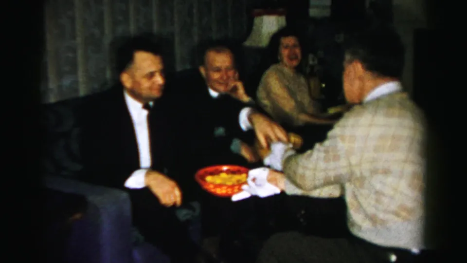 1956 Albany party frame with tuxedoed men, a woman in the background, and a gloved man presenting a red snack bowl.