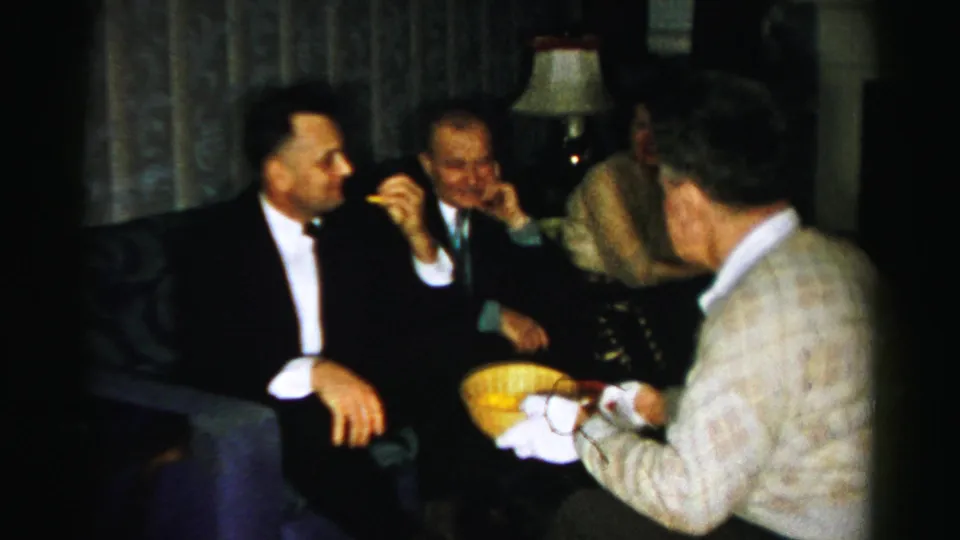 Albany, New York 1956 home-party frame showing a gloved man offering a yellow snack bowl to guests in formal dress.