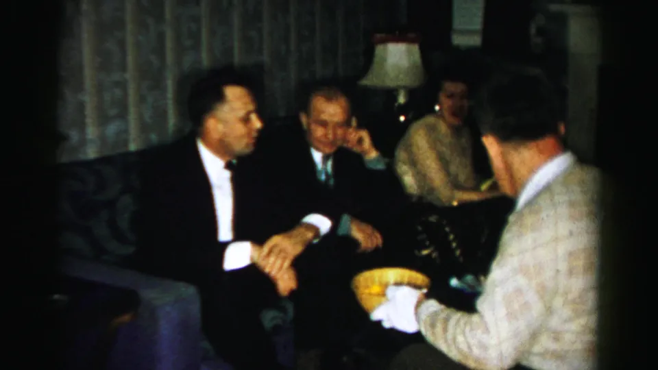 1956 Albany indoor party frame with tuxedoed guests and a gloved man holding a yellow snack bowl in a private living room.