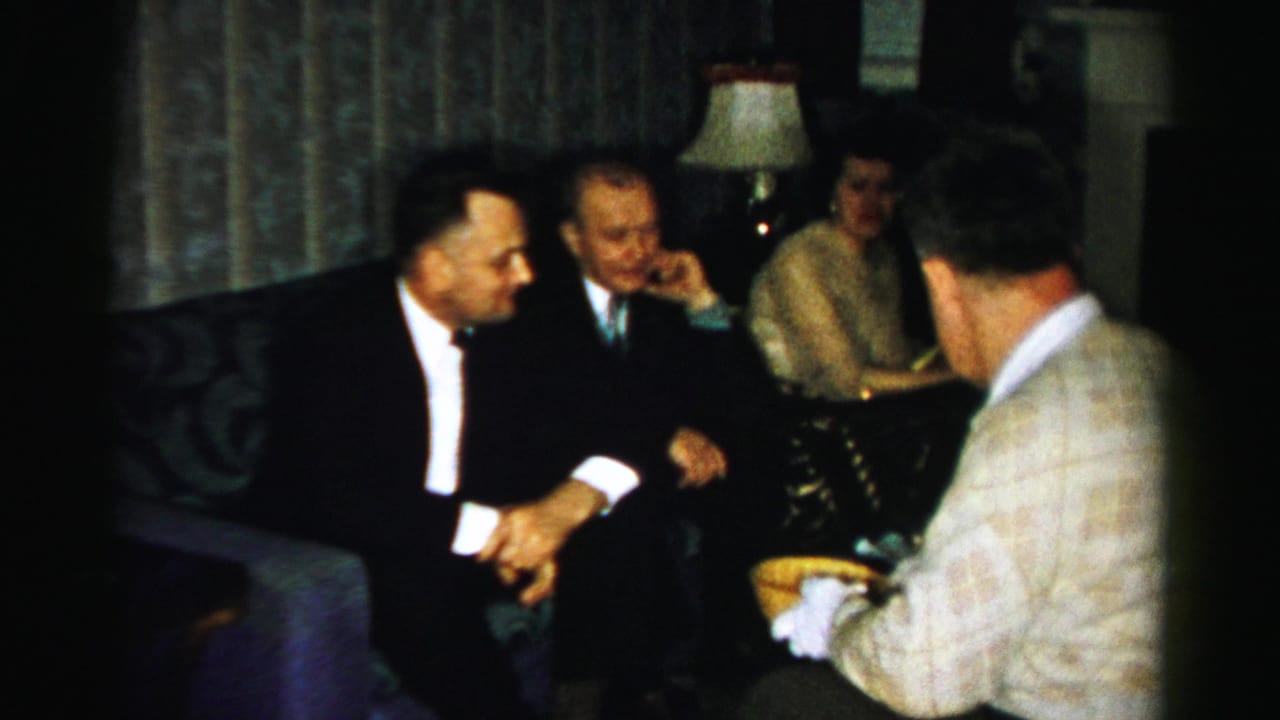 Adults in a dim living room sit and talk while a man in a patterned sweater serves food.