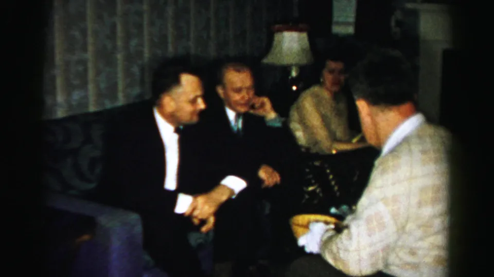 In Albany home-movie footage from the 1950s, suited men and a seated woman gather in a dim living room as food is served.