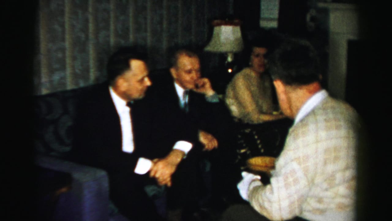 A 1950s Albany living-room gathering shows suited men in conversation while a man with his back to camera holds a serving bowl.