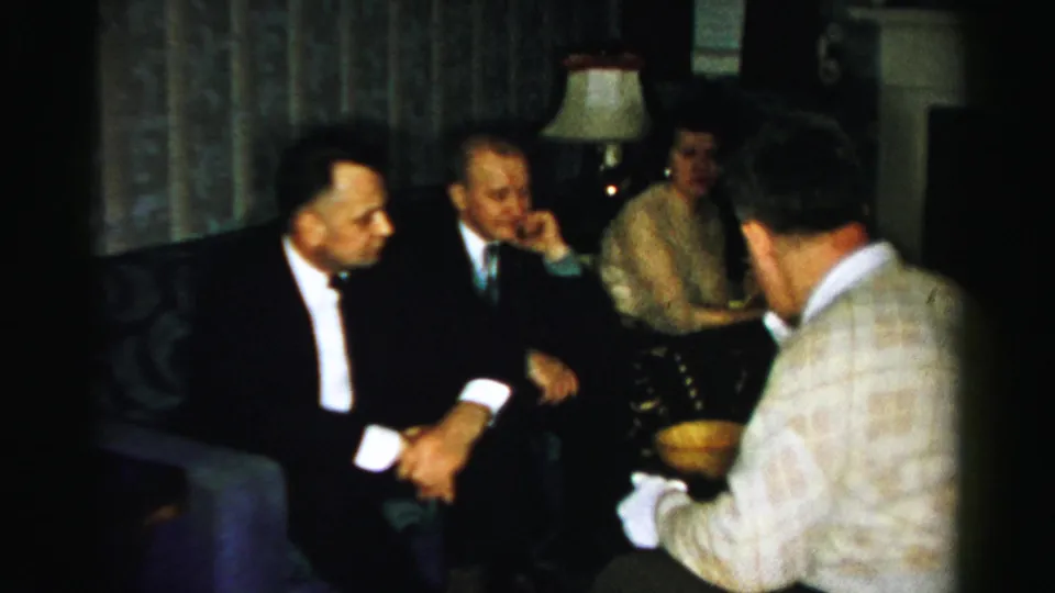 Suited men and a woman sit closely in a private Albany interior, with a sweatered man serving food in 1950s home-movie footage.
